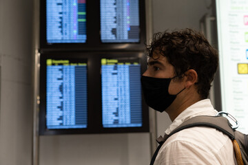 Fototapeta na wymiar Young traveler man with brown hair and brown eyes carefully observes the screens of the flights at the airport, wears a white shirt with a backpack and wears a Mask against Covid19