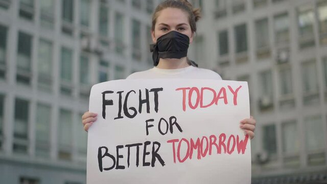 Beautiful woman in face mask standing in urban city with banner and looking at camera. Portrait of female Caucasian demonstrator protesting against social inequality and rights violation.