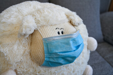Close-up of a fat stuffed sheep with a face covering mask. This plush animal is in quarantine at home due to the Covid-19 pandemic.