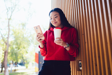 Attractive female influence blogger checking popularity of own web blog using 4g wireless connection on modern cellular gadget, pretty Japanese woman with coffee to go enjoying online chatting