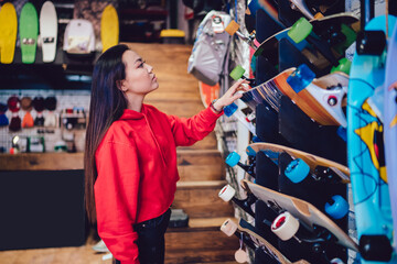 Side view of attractive teenage woman choosing longboard from store rack during Black Friday sales, beautiful Japanese hipster girl spending day for buying skateboard and practicing hobby skills
