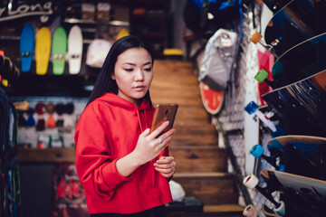 Fototapeta na wymiar Millennial female teenager using cellphone for browsing web store and compare prices during shopping day for choosing youthful skateboard, Chinese generation Z making banking payment for purchase