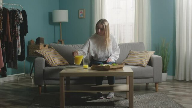 Young girl in white jacket sits on beige sofa and pours orange juice into glass. The camera hits the subject