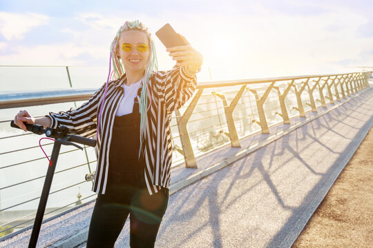 Girl with dreadlocks is standing on bridge, making selfie on smartphone. Young woman in yellow glasses came to meeting by electric scooter. Moving by ecological eco portable transport of future.