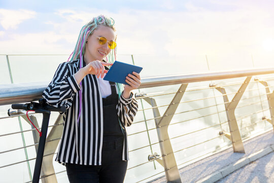Girl with colored dreadlocks is standing on bridge, holding tablet. Young woman in yellow glasses came to meeting by electric scooter. Moving by ecological eco portable transport of future concept.