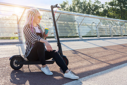 Portrait of girl with colorful dreadlocks is sitting on electric scooter on bridge in city. Young woman in yellow glasses is drinking coffee. Moving by ecological eco portable transport of future.