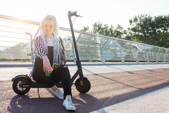 Portrait of girl with colorful dreadlocks is sitting on electric scooter on bridge in city. Young woman in yellow glasses is drinking coffee. Moving by ecological eco portable transport of future.