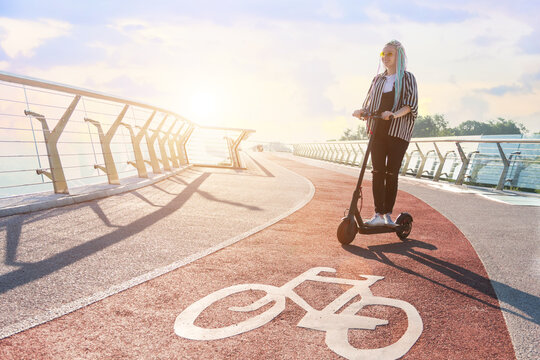 Smiling girl with colorful dreadlocks is riding, driving electric scooter on bike path. Young woman in yellow glasses is walking on bridge of modern city. Moving by ecological eco portable transport.
