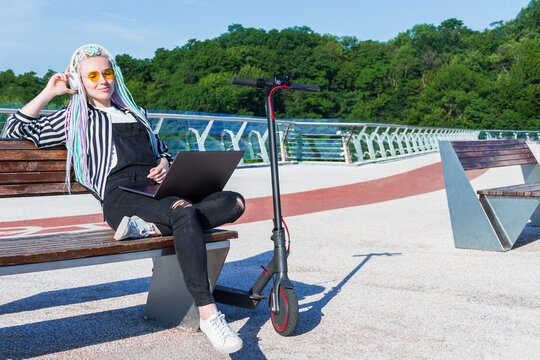 Girl with colored dreadlocks is working on laptop, listening to music outdoor. Young woman freelancer, designer in yellow glasses is sitting on bench in modern city. Moving by electric scooter concept - Powered by Adobe