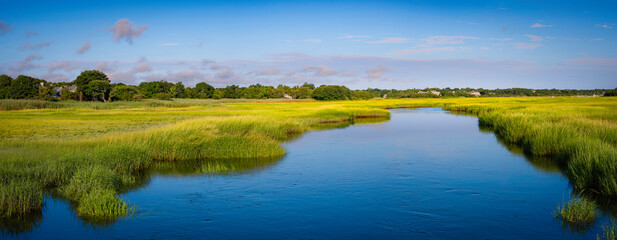 Panoramic landscape of wide blue waterway through green marsh field with views of trees and clouds