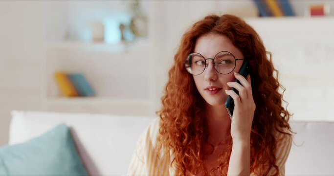 Pretty young woman with red curly hair having pleasant conversation on mobile phone smiling laughing feeling excited nodding head in agreement. Portrait of happy lady talking on cellphone on sofa.