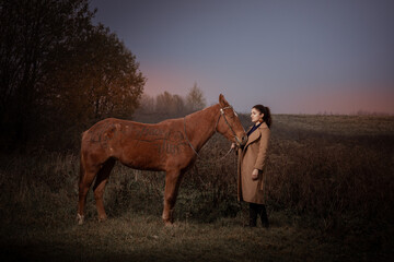 Fototapeta na wymiar Young brunette woman in a beige coat posing with a red brown horse. Sunset sky and autumn nature. The inscription on the horse: have fun.