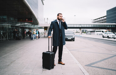 Handsome man with suitcase talking on phone near airport