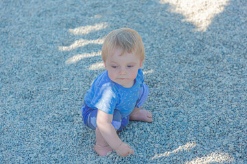 Fototapeta premium Funny baby sitting on the ground, covered with small stones. A blond boy plays with pebbles in a city Park.
