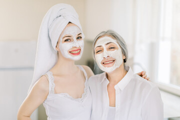 Portrait of two Caucasian women, young girl with hair wrapped in towel, and elderly gray haired woman, with homemade facial masks on faces, smiling to camera, posing at home. Beauty treatment