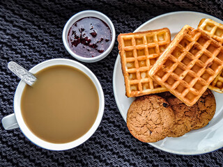 Homemade cakes, instant coffee with milk, raspberry jam on a black knitted background. Waffles and oatmeal cookies made at home. Homemade food and drink close-up.