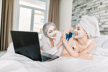 Two women with facial masks, pretty young girl wrapped in towel and her senior attractive grandmother, having fun while watching movie on laptop at home, and giving high five each other