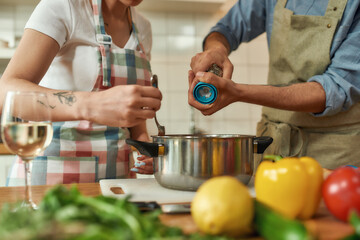 Cropped shot of man adding pepper, spice to the soup while woman stirring it with a spoon. Couple preparing a meal together in the kitchen. Cooking at home, Italian cuisine