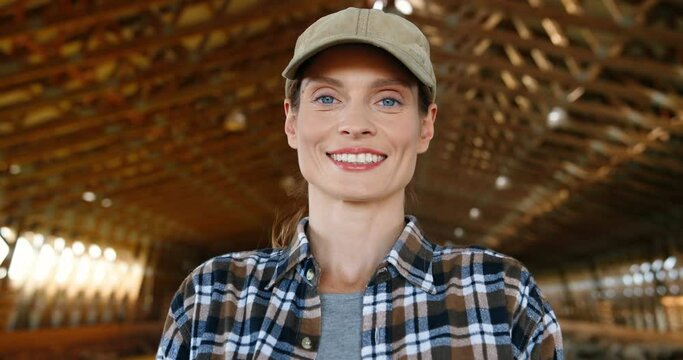 Young beautiful Caucasian woman farmer looking at camera and smiling in barn with livestock. Portrait of happy cheerful female shepherd with smile in stable. Indoors. Dolly shot. Zooming in face.