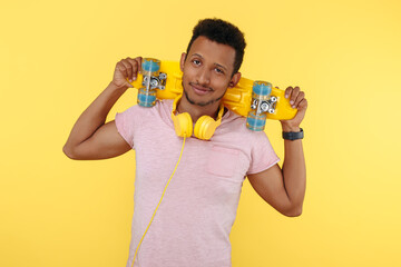 Hesitating african american young man wearing headphones, holding skateboard on shoulders over yellow background.