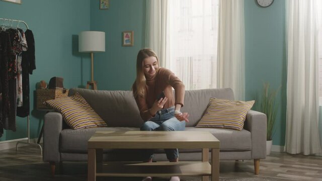 Young girl in brown sweater and jeans sits on beige sofa and takes picture of hamster on her mobile phone. Camera hits subject.