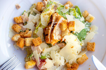 Top view of fresh healthy Caesar salad with grilled chicken breast in a big white plate with fork and knife aside.