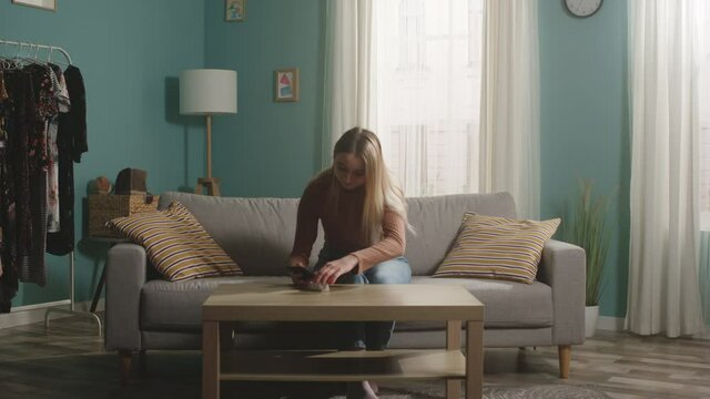 Young girl in brown sweater and jeans sits on beige sofa and takes picture of hamster on her mobile phone. Camera hits the subject.