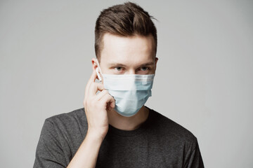 a young man in a mask looks at the gadget with a surprised face. grey t-shirt and background. a protective agent against the pandemic face. fashionable hairstyle. presses a finger on the phone