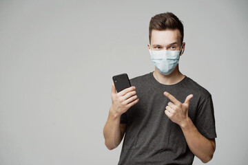 portrait of a young man looking at a friend, wearing a mask and pointing a finger at the phone. grey t-shirt, grey background. protection from pandemic face