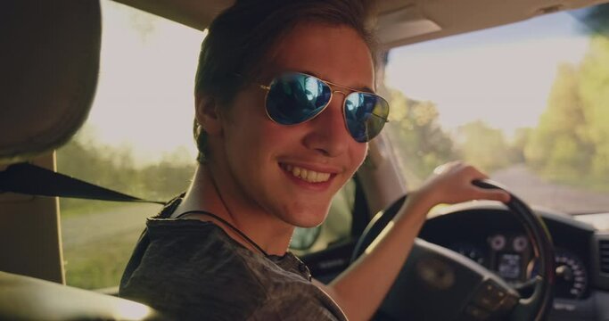 Young happy guy in the car looking to the camera. Handsome young smiling man in sunglasses is sitting in a car at sunset. Sun's rays shine through the windshield of the car.
