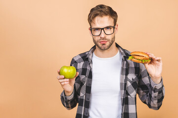 Young man holding green apple and a hamburger isolated over beige background. Concept of choosing between healthy and unhealthy foods.