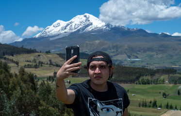 
man taking a selfie in the green field with a snowy volcano in the background with blue sky and some clouds 