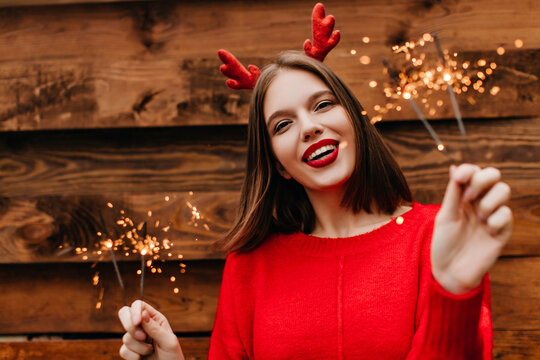 Lovely female model with straight hairstyle holding bengal lights with inspired smile. Cute woman in red sweater preapring for new year.