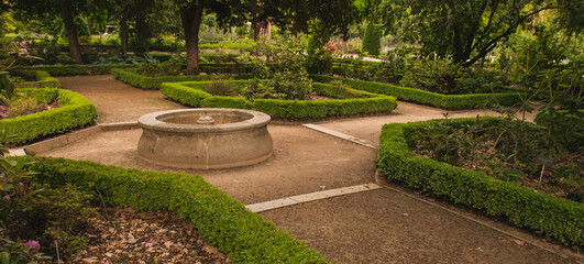 Obraz premium Fuente redonda de piedra situada en un cruce de caminos dentro de un jardín botánico, rodeada de parterres con diferentes tipos de plantas y árboles.