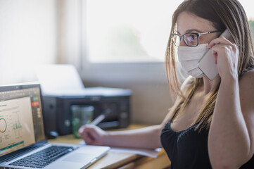 Mujer ejecutiva con gafas trabajando hace una llamada profesional con teléfono móvil y habla con una mascarilla higiénica puesta desde su puesto de trabajo.