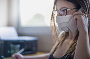 Mujer ejecutiva con gafas trabajando hace una llamada profesional con teléfono móvil y habla con una mascarilla higiénica puesta desde su puesto de trabajo.