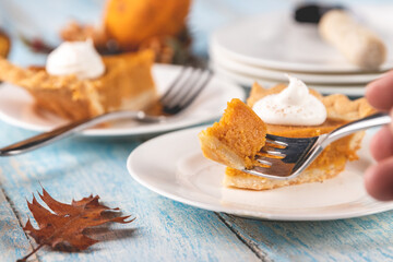 Closeup perspective shot of a bite of pumpkin pie on a fork with plates and slices of pumpkin pie blurred in the background on a rustic blue painted wood table surface.