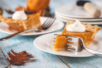 Closeup perspective shot of a slice of pumpkin pie with a fork cutting into it with plates and pie blurred into the background on a rustic blue painted wood table surface.