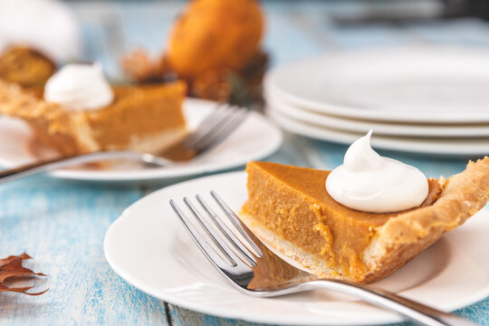 Closeup perspective shot of a slice of pumpkin pie on a plate with a fork with plates and pie blurred in the background on a rustic blue painted wood table surface.