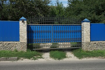 large blue iron gate with a black forged pattern and part of the wall of the fence made of metal and gray stones on the street