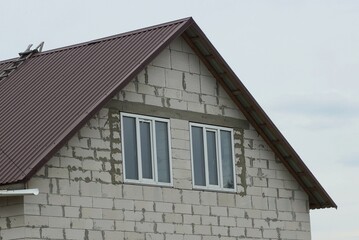 attic of a white brick house with two windows under a brown tiled roof against a gray sky