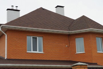 red brick house with white windows and brown tiled roof with chimneys against gray sky