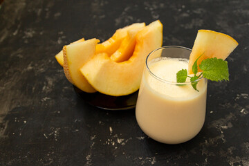 Melon milk shake on a black background. Ripe sliced melon is lying nearby. The smoothie glass is decorated with a sprig of mint and melon crumbs. Summer healthy food concept