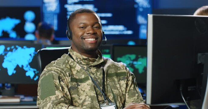 Portrait of African American male army officer in uniform sitting at desk, working on computer in analytic secret base and smiling to camera. Man militarian official in dark office. Digital support.