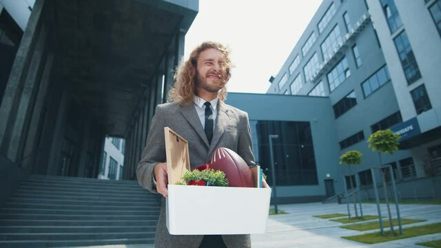 Handsome curly-haired fired man with personal box feel happy to quit job leaving office department smiling very satisfied.
