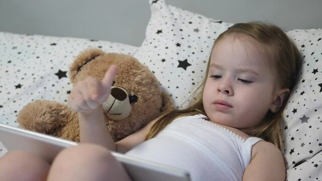 Happy child is playing a tablet. A little girl lies in bed with a teddy bear.
