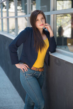 Latin white skinned woman with long hair, posing standing by a window, is wearing a yellow blouse, blue jacket, jeans and belt, photo in the day