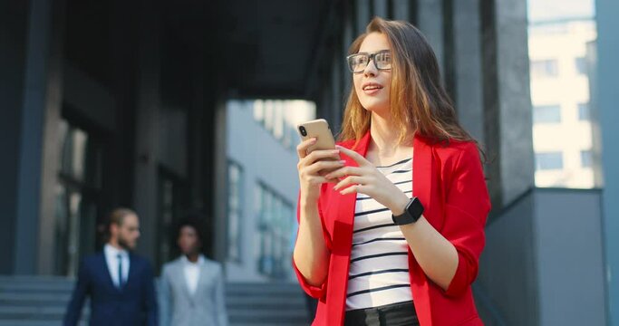 Pretty young smiled Caucasian woman in red jacket smiling and texting message on mobile phone at street. Outdoors. Cheerful female tapping and scrolling on smartphone. Girl chatting in social media.