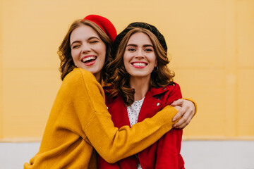 Glad white woman with curly hairstyle embracing her brunette sister outdoor. Portrait of refined female models expressing happiness on yellow background.