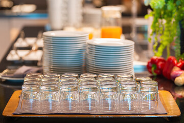 A set of empty small glasses for fresh juice and Group of white plates stacked together. Blurred background breakfast buffet line in hotel.